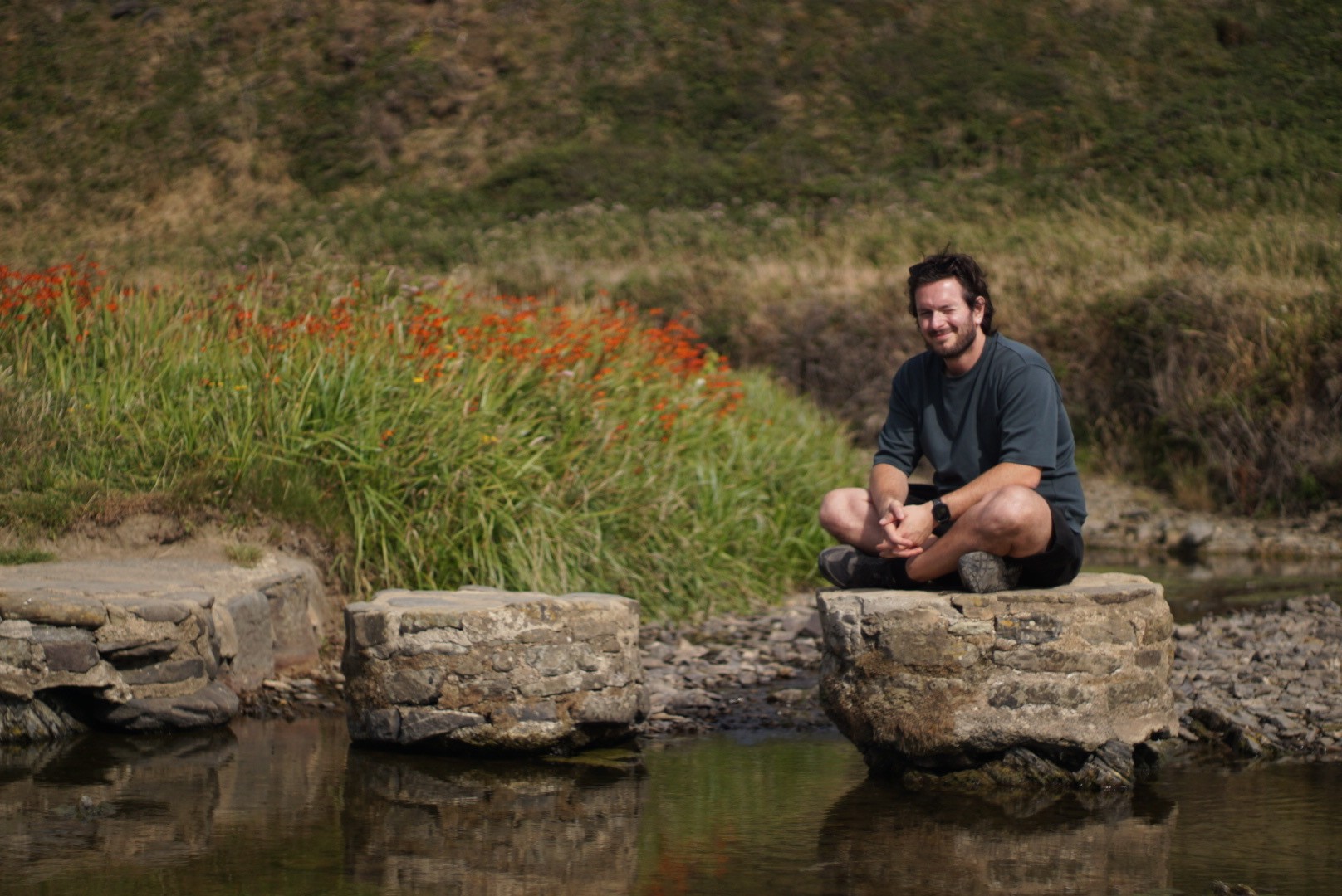 Domenico sitting cross-legged on a stepping stone in a stream, with orange flowers and a grassy bank behind him